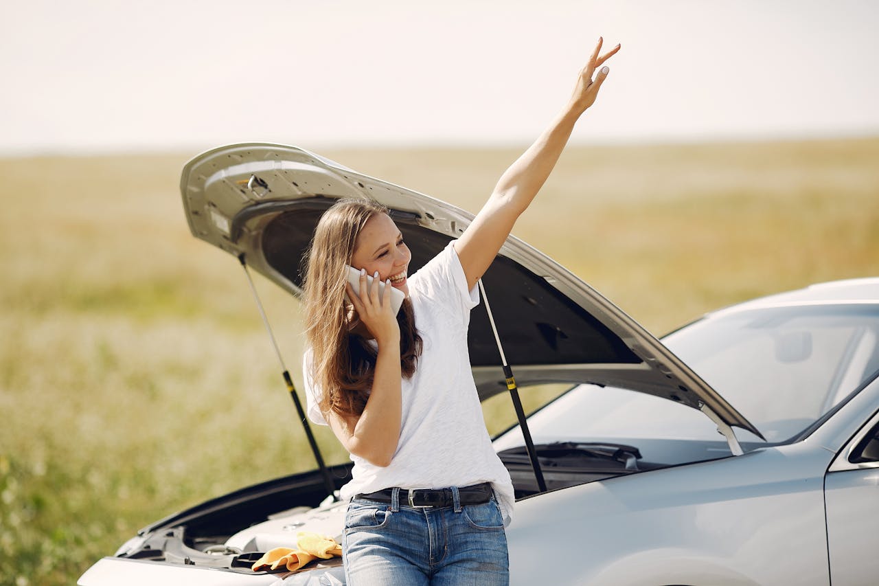 Services-02 Cheerful young female driver in white t shirt and jeans speaking on phone and looking away with smile while leaning on broken car with open hood and showing greeting gesture with raised hand
